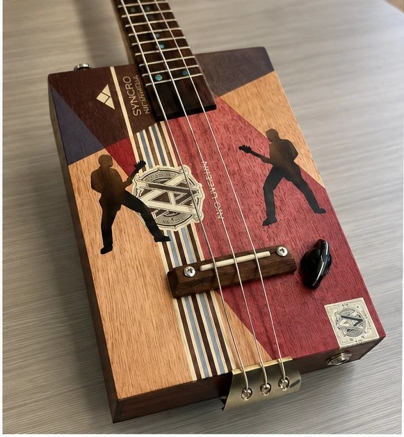 Top-down view of the cigar box guitar's body, painted with geometric color blocks and silhouettes of guitarists, with a wooden bridge and brass tailpiece