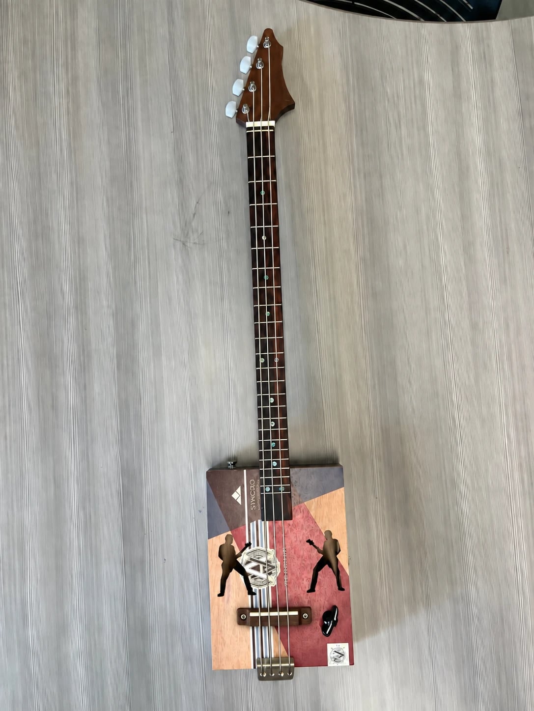 The finished cigar box guitar lying on a light wood-grain table, seen from above: a long dark fretboard with shell position markers and a pointed wooden headstock with four tuning pegs, leading down to a cigar-box body inlaid with geometric blocks of color and two guitarist silhouettes, with a wooden bridge and brass tailpiece