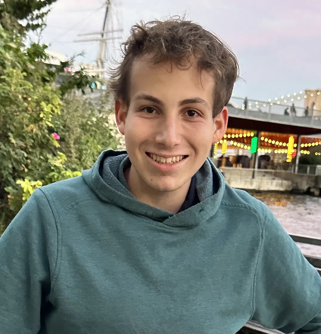 Noah Spiegelman smiling outdoors in a teal hoodie, with a waterfront behind him
