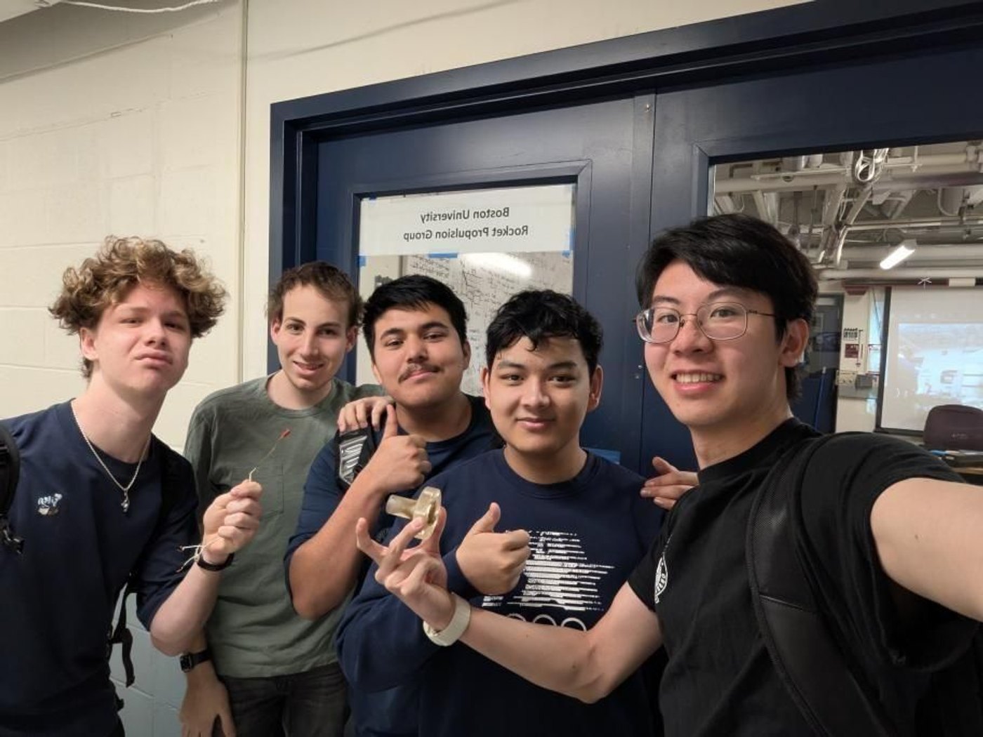 Five team members posing in front of the Boston University Rocket Propulsion Group door, one holding up the nozzle