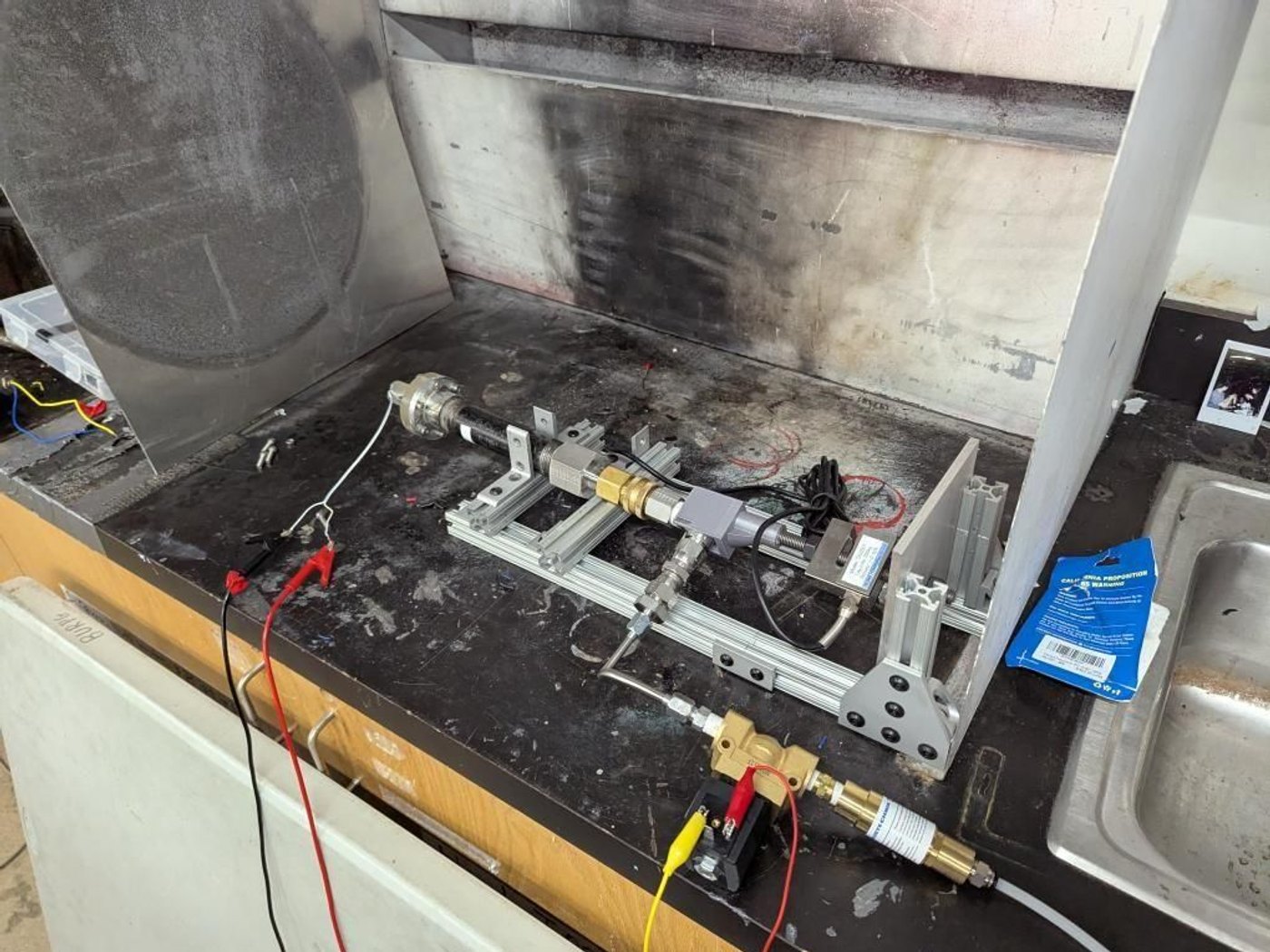 The test stand inside a fume hood before the firing: an aluminum-extrusion frame holding the chamber and nozzle, a load cell, brass valve fittings and wiring