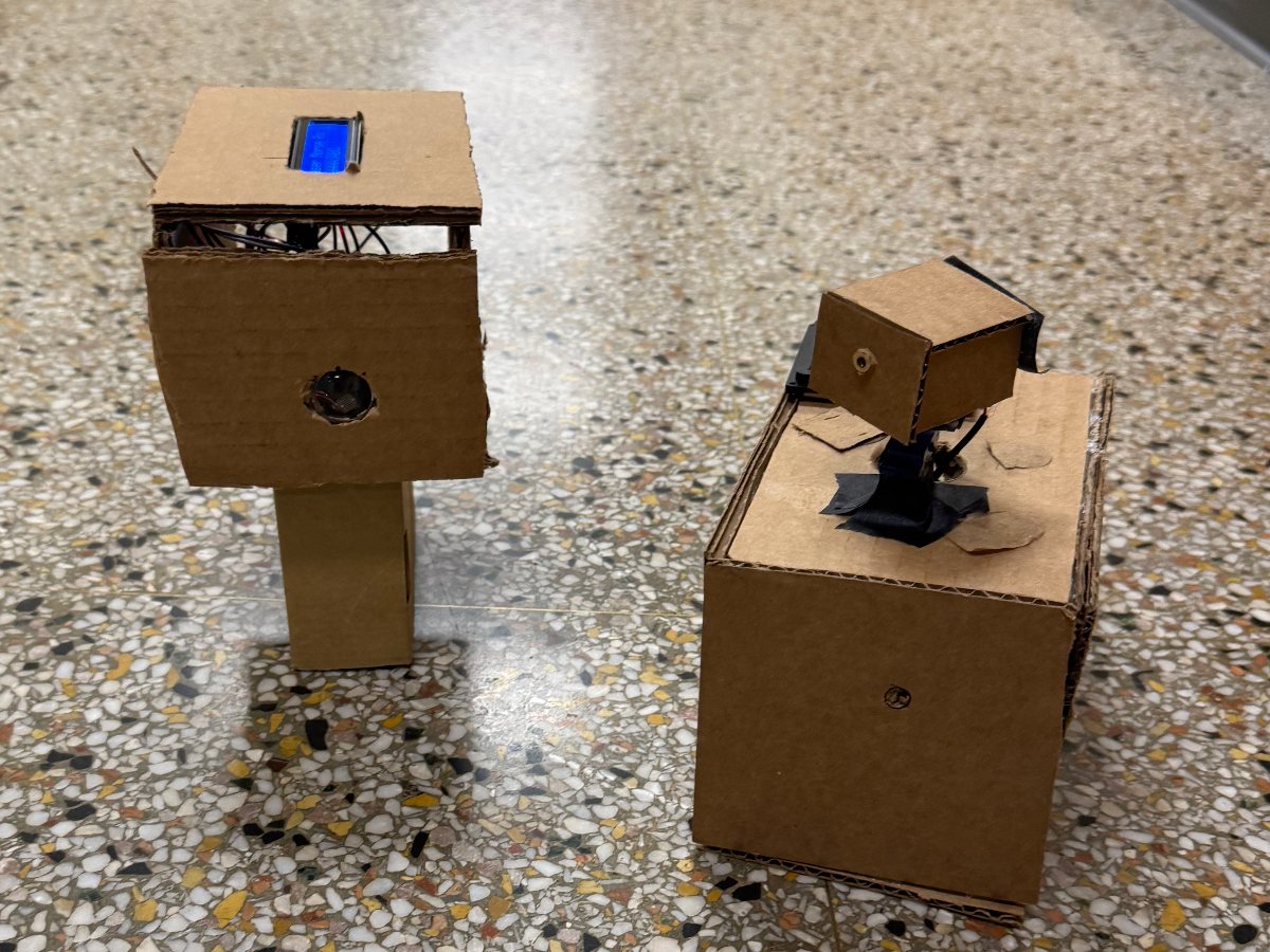 Two cardboard prototype units on a hallway floor: the receiver on the left with a glowing blue LCD on top, and the transmitter on the right with a small servo-mounted laser housing on its lid