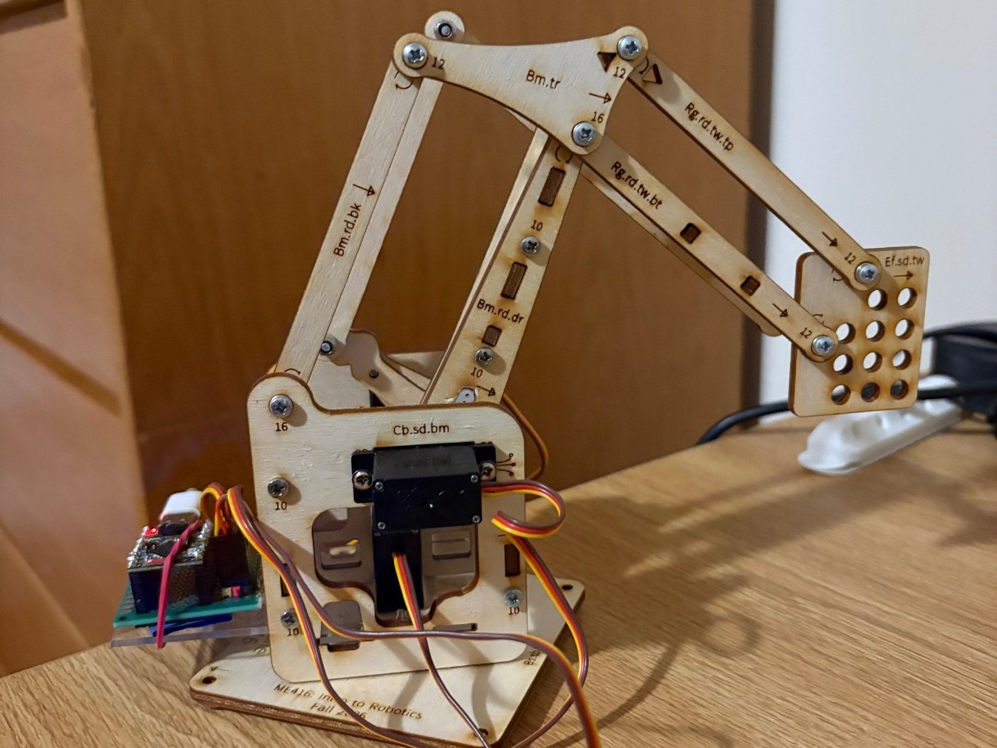 Angled view of the robotic arm reaching to the right, with parallel wooden links held together by screws and the base engraved with ME416: Intro to Robotics, Fall 2026