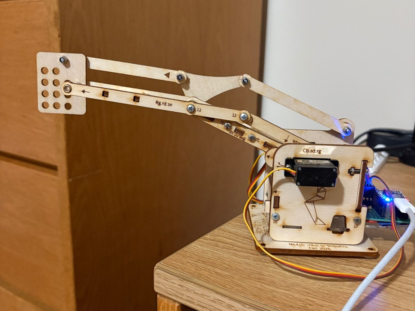 Side view of the wooden robotic arm reaching out to the left from its base, with three servo wires and a USB cable running to the ESP32 board on the right