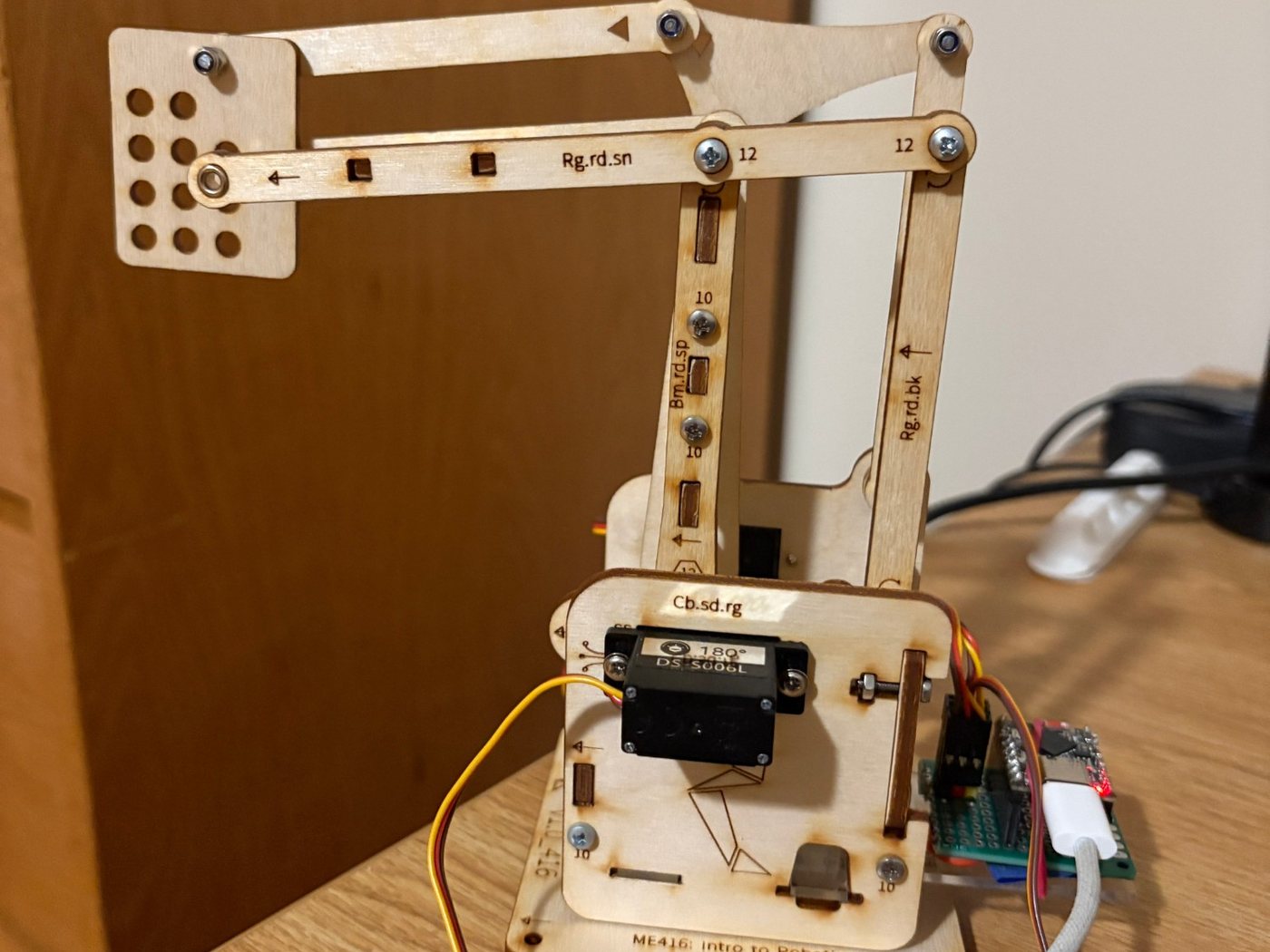 Front view of the robotic arm's base with its servo, laser-engraved labels and the soldered ESP32 board, and the arm extended behind it