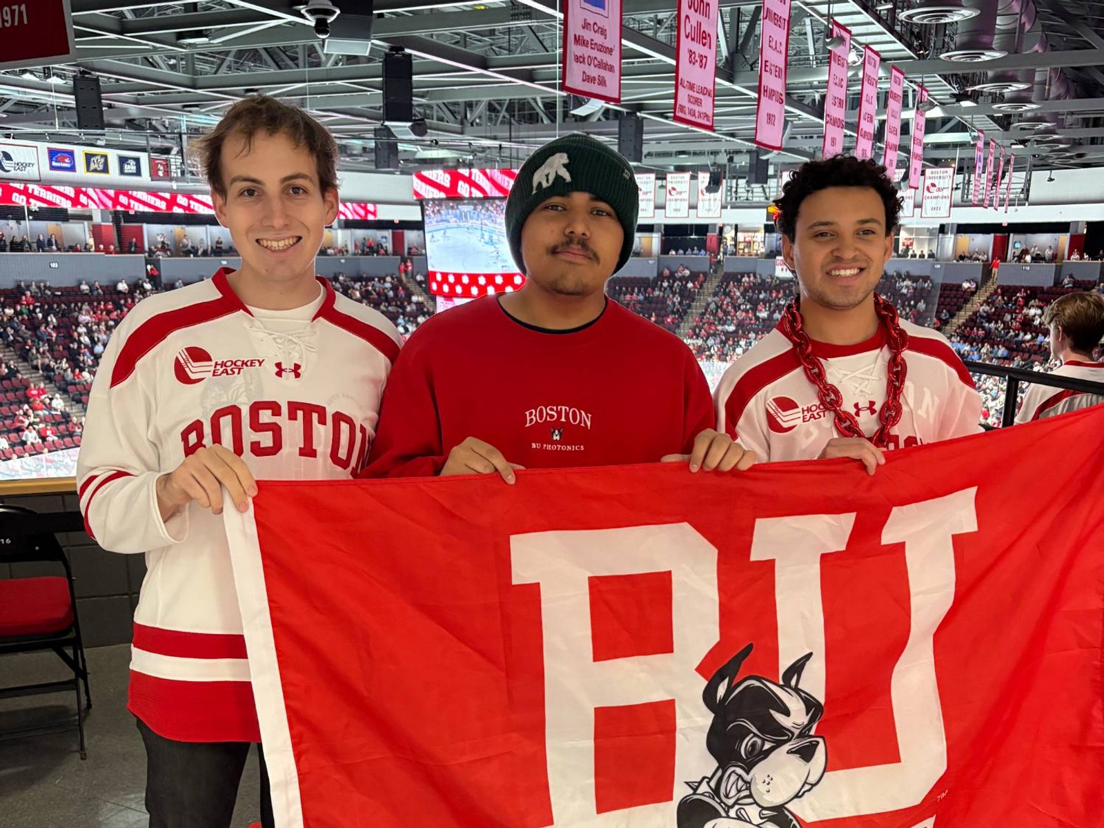Three RASTIC teammates smiling at a Boston University hockey game, holding up a red BU flag