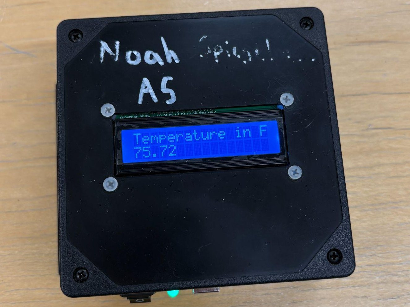 The finished black enclosed temperature monitor on a table, with a blue 16 by 2 LCD reading Temperature in F, 75.72, and a green power LED lit at the edge