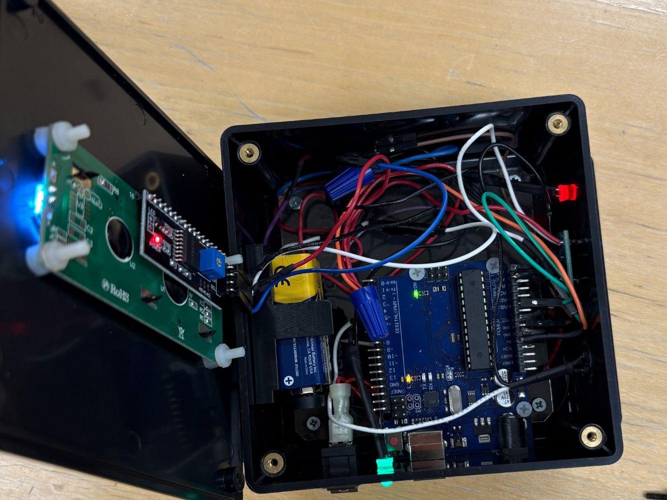 The monitor with its lid open, showing the Arduino Uno, a 9 V battery in a 3D-printed holder, twist-nut caps, colored wiring and the red LED