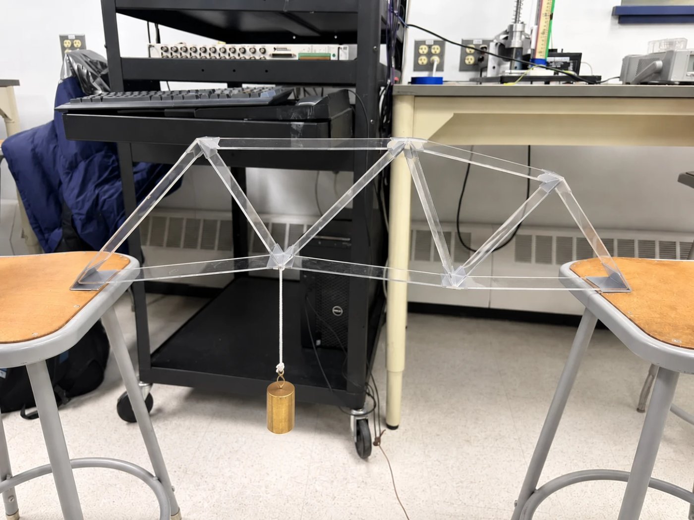 The physical acrylic truss resting on two lab stools during load testing, with a weight hanging from its center joint